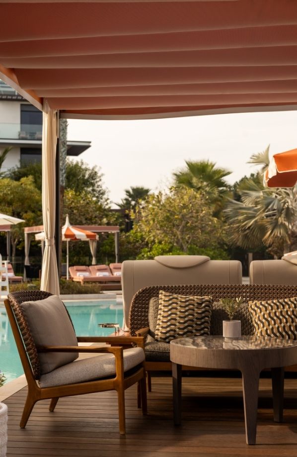 Luxurious outdoor lounge area beside a swimming pool, featuring stylish wooden and woven furniture with plush cushions. A shaded pergola with fabric draping provides comfort, while patterned pillows and a modern coffee table enhance the elegant ambiance. Sun loungers and cabanas with striped umbrellas are visible in the background, surrounded by lush greenery